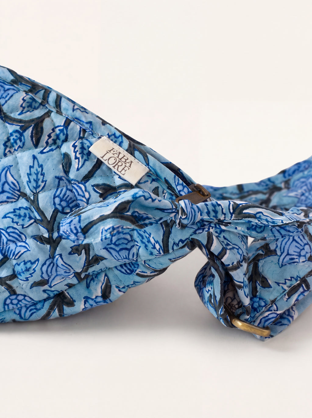 Closeup of Fabalore Blue Bloom sling bag showing floral fabric, zipper, logo label and strap hardware.