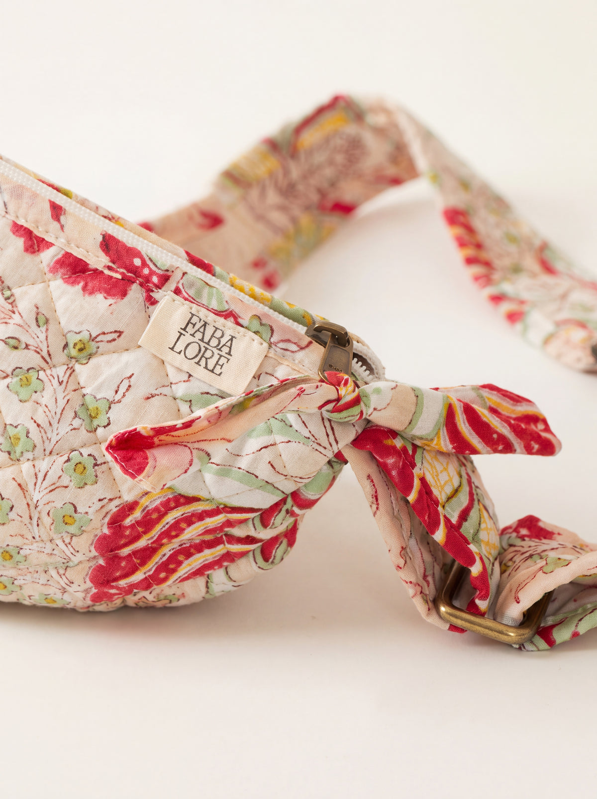 Close up of Fabalore Meadow quilted sling bag showing floral fabric, zipper, logo label and adjustable strap hardware.